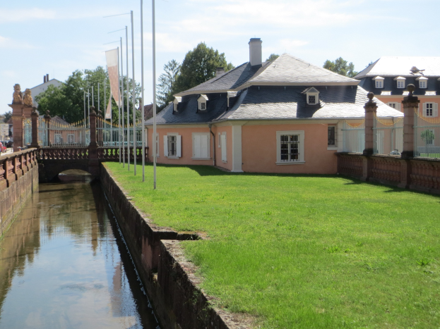 Kleiner Kanal mit einer Brücke, der durch ein grünes Feld neben einem Gebäude läuft, unter einem klaren blauen Himmel mit Bäumen und Gebäuden im Hintergrund.