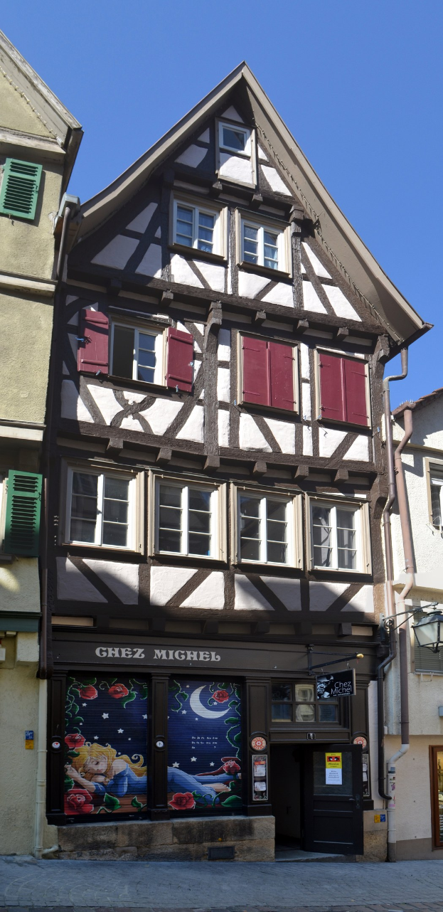 Altes Gebäude mit roten Fensterläden an einer Straße, mit mehreren Fenstern und einem klaren blauen Himmel im Hintergrund.