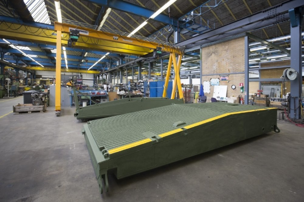 Großes Lagerhaus mit grünen Containern, Maschinen, Polen, Lampen, Stangen und einer Wand mit einer Uhr und Papieren.