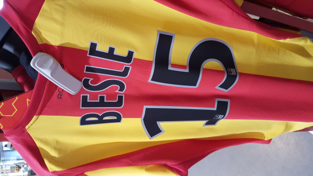 Ein rotes und gelbes Fußballtrikot mit aufgedrucktem Text und Nummern, das an einer Stange in einem Geschäft hängt, mit anderen Gegenständen und dem Boden im Hintergrund.