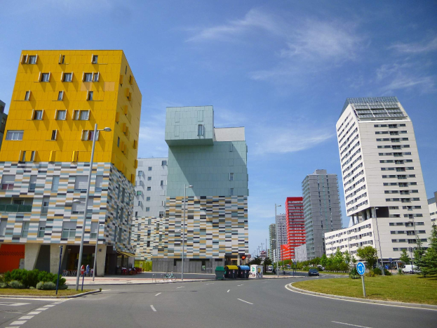 Eine Stadtstraße mit hohen Gebäuden im Hintergrund, Straßenlaternen, Straßenlaternen, Kraftfahrzeugen, Infotafeln, Büschen, Bäumen und einem bewölkten Himmel, mit einzigartig gestalteten Gebäuden der Universität Rotterdam.