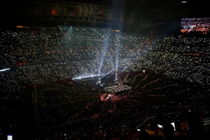 Eine gro├če Menge Menschen in einem Stadion bei Nacht, beleuchtet von hellen Lichtern, mit einem Bildschirm auf der rechten Seite, der die Ergebnisse der Super Bowl XLVII-Halbzeitenshow anzeigt.