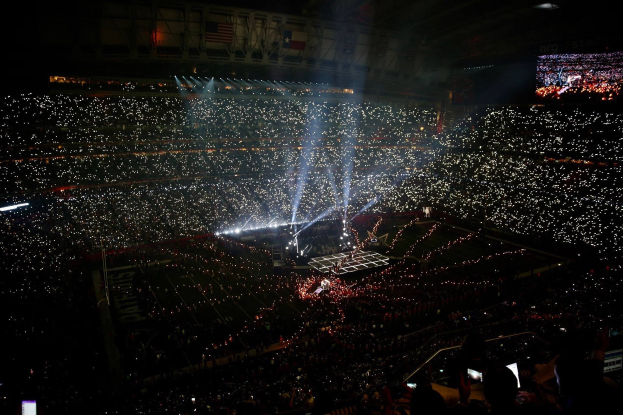 Eine gro├če Menge Menschen in einem Stadion bei Nacht, beleuchtet von hellen Lichtern, mit einem Bildschirm auf der rechten Seite, der die Ergebnisse der Super Bowl XLVII-Halbzeitenshow anzeigt.