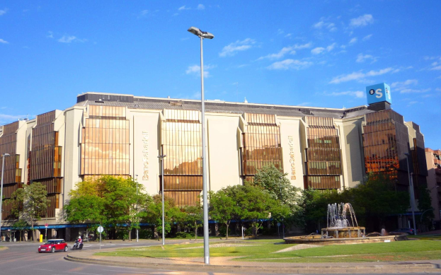Großes Gebäude namens Bancroft-Stadion mit einem Brunnen davor, umgeben von Straßenlaternen, Lampen, Fahrzeugen, Passanten, Bäumen und einem bewölkten Himmel.