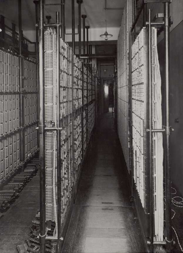 Schwarzes und weißes Foto eines langen Datenzentrumsgangs mit Metallrohren, Deckenleuchten und Reihen von Servern.