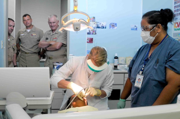 Eine Gruppe von Menschen in einer Zahnarztpraxis mit einem Zahnarzt in einem weißen Kleid, der ein Objekt hält, einer weiteren Person mit Brille, Mundschutz und einem Ausweis, Plakate an der Wand und eine Tür auf der linken Seite.