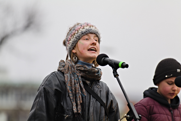 Eine Frau in schwarzer Jacke, Schal und Mütze singt in ein Mikrofon auf der Bühne, neben einer Person in einer maronenfarbenen Jacke und schwarzer Mütze, während der unscharfe Hintergrund einen träumerischen Effekt schafft.