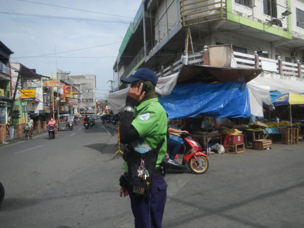Ein Polizeibeamter in grüner und blauer Uniform und blauer Mütze steht an der Straße und spricht in ein Handy, mit Fahrzeugen im Hintergrund, Ständen rechts und einem bewölkten Himmel.