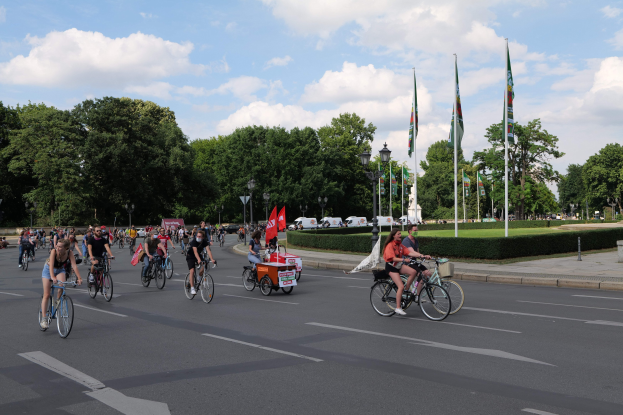 Eine Gruppe von Menschen, die auf Fahrrädern eine von Bäumen gesäumte Straße mit Laternenmasten und Fahnenmasten entlangfahren, während des Pariser Radrennens 2016, mit bewölktem Himmel und im Hintergrund sichtbaren Fahrzeugen.