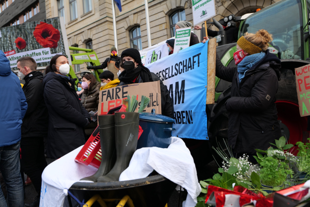 Eine Gruppe von Menschen mit Masken hält Protestschilder vor einem Lastwagen, mit einem Tisch, Pflanzen, Gebäuden und einer Flagge im Hintergrund, versammelt gegen ein Regierungsverbot von Pestiziden.