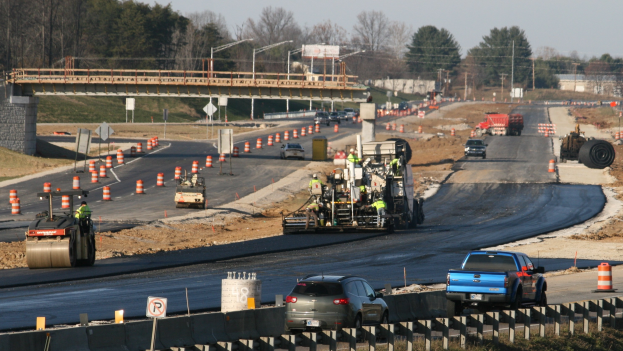 Baustelle mit Fahrzeugen auf der Straße, Verkehrsleitkegel, Hinweisschilder, Versorgungsmasten, eine Brücke, Bäume und einen klaren blauen Himmel.
