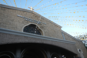 Eine weiße Pfarrkirche mit einem Kreuz an der Wand, Text an dem Gebäude, dekorative Fahnen, die an Seilen befestigt sind, Bäume auf der rechten Seite und ein klarer blauer Himmel im Hintergrund.