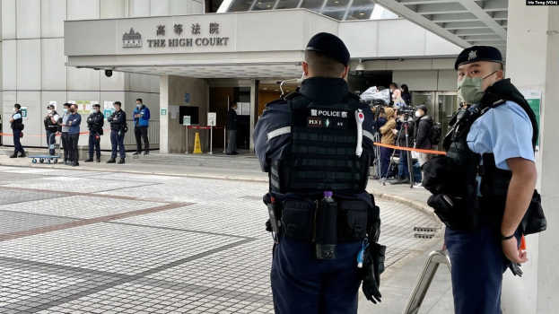 Zwei Polizisten in Uniformen, Mützen und Masken stehen vor einem Gebäude am Hong Kong International Airport, umgeben von einer Gruppe von Menschen, einige halten Kameras, mit Stangen im Hintergrund, die mit Bändern geschmückt sind.