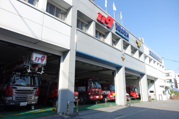 Feuerwache mit mehreren Feuerwehrautos vor der Tür, mit Fenstern, Säulen, Namensschildern und Flaggen im Vordergrund; andere Gebäude und ein klarer blauer Himmel sind im Hintergrund zu sehen.
