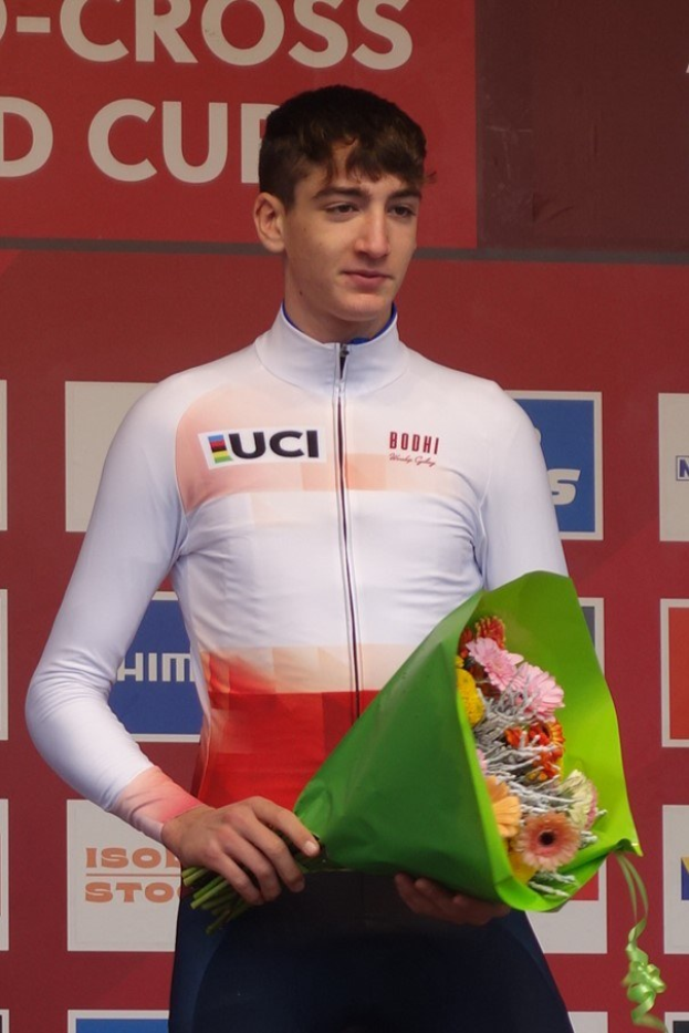 Ein Mann steht auf einem Podium und h├Ąlt einen Blumenstrau├če, feiernd mit einem selbstbewussten und stolzen Gesichtsausdruck.