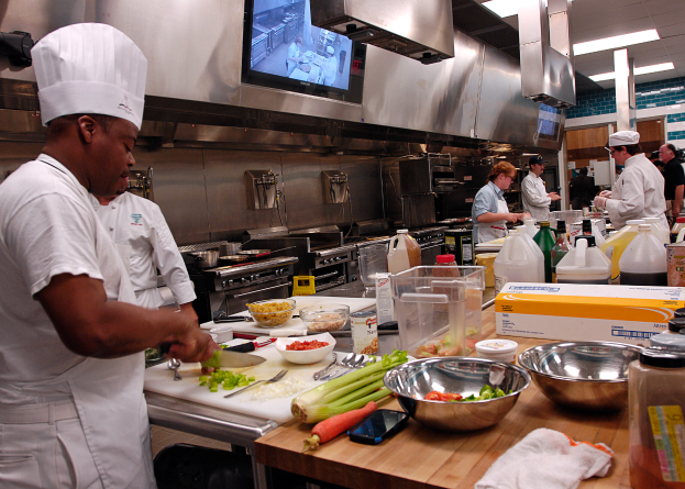 Eine Gruppe von Köchen in einer Küche, die auf einem Schneidbrett Essen zubereiten, mit dem Koch im Vordergrund in einer weißen Schürze und Mütze, der ein Messer hält, und umgeben von Utensilien, Schüsseln, Gemüse, einem Mobiltelefon, Flaschen und anderen Gegenständen, mit einem Fernseher und Deckenlampen im Hintergrund.