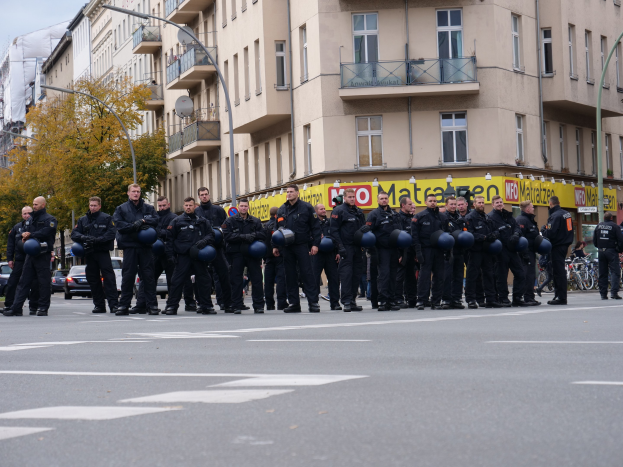 Polizisten in schwarzen Uniformen mit blauen Helmen stehen in einer Straße, die von Laternenmästen, Bäumen und glasfensterigen Gebäuden gesäumt ist, unter einem klaren blauen Himmel.