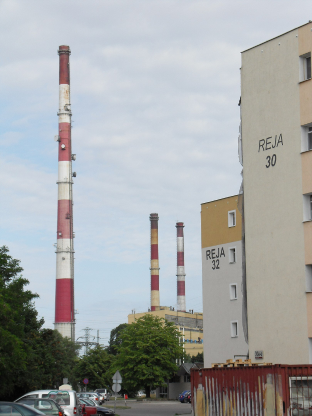 Eine Fabrik mit parkenden Autos vorne, umgeben von Gebäuden, Bäumen, Strommasten, Schildern und einem Container unter einem bewölkten Himmel.