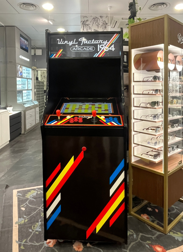 Ein vintage Arcade-Automat steht in der Mitte eines retro-stylischen Ladens, mit einem Gestell mit Gläsern rechts und Schränken, einem Fernseher und anderen Gegenständen im Hintergrund unter Deckenbeleuchtung.