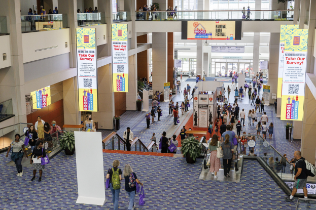 Eine große Menschenmenge, die durch ein großes Gebäude mit Bannern, Topfpflanzen, Rolltreppen, Geländern, Säulen, bedruckten Schildern, Glastüren und anderen Gegenständen geht, wobei einige Taschen tragen, beim Nationalen Buchfestival.