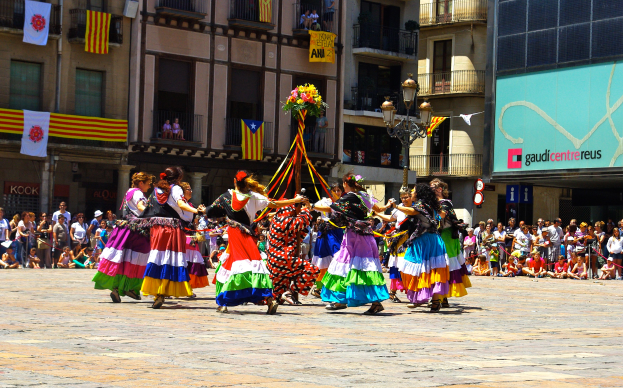 Eine Gruppe von Frauen in bunten Kleidern tanzt auf der Straße, umgeben von einer Menge Menschen, mit Gebäuden, Geländern, Balkonen, Fahnen, einem Laternenpfahl und einer Tafel mit Schrift im Hintergrund.