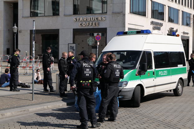 Eine Gruppe von Polizisten steht vor einem Starbucks-Café, rechts daneben ein Van und links ein paar Menschen; im Hintergrund sind ein Gebäude, ein Schild, ein Lichtmast und ein Zaun zu sehen.
