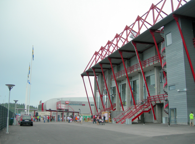 Großes Stadion mit Menschen, die herumlaufen, Fahrzeugen auf der Straße davor, einem Zaun auf der linken Seite, Bäumen und einem klaren blauen Himmel im Hintergrund.