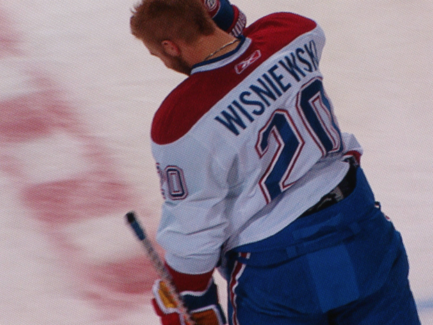 Ein Hockey-Spieler in einem 'Wisniewski'-Trikot steht auf dem Eis und hält einen Hockey-Schläger, mit konzentrierter und entschlossener Miene.