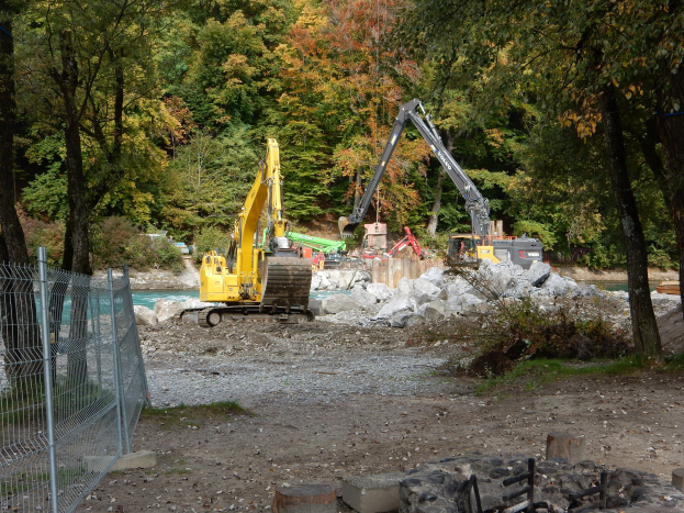 Baustelle mit Kränen, Steinhaufen, Holzstämmen, Vegetation, einem Metallzaun, Bäumen, einem Gewässer und einem bewölkten Himmel.