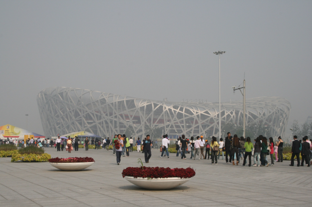 Olympisches Stadion in Beijing mit Menschen, die herumlaufen, Pflanzen mit Blumen im Vordergrund, Laternenmasten, Bäumen und einem klaren blauen Himmel im Hintergrund.