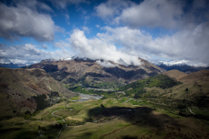 Panoramischer Blick von der Spitze eines Berges in Queenstown, Neuseeland, mit saftigem Grün, verstreuten Bäumen und einer gewundenen Straße unter einem Himmel voller weißer, flauschiger Wolken.