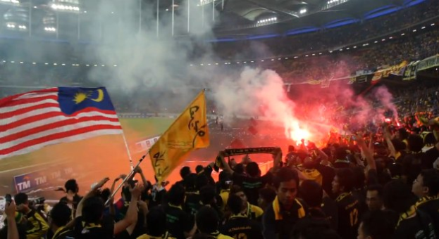 Eine große Menschenmenge in einem Stadion mit Raketen, Fahnen und einer Kamera, mit Rauch und Schildern mit der Aufschrift "Malaysia vs Malaysia Live Streaming" im Hintergrund.