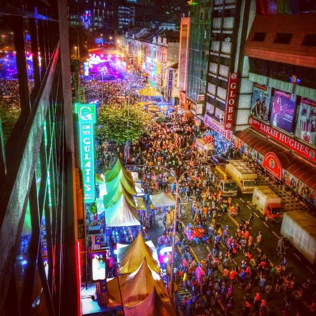 Eine volle Faschings-Szene mit Zelten, Geschäften, Gebäuden und Bannern, die die Straße säumen, mit hellen Lichtern im Hintergrund.