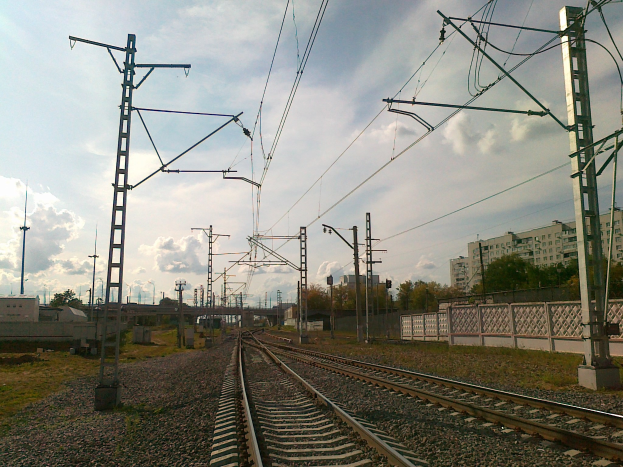 Eisenbahnschiene mit Oberleitungen, umgeben von Steinen, Gras, einem Zaun, Bäumen, Strommasten, einer Brücke, Gebäuden und einem bewölkten Himmel.