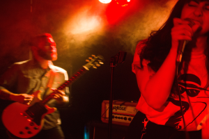 Eine Frau singt in ein Mikrofon auf der Bühne, während ein Mann im Hintergrund Gitarre spielt und Deckenlampen das lebendige Setting beleuchten.