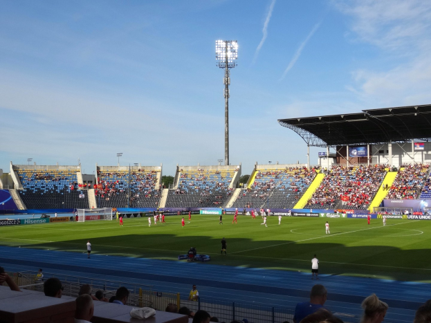 Ein Fußballspiel in einem großen Stadion mit Spielern auf dem Feld und Zuschauern in den Rängen unter Flutlicht, mit dem Himmel im Hintergrund.