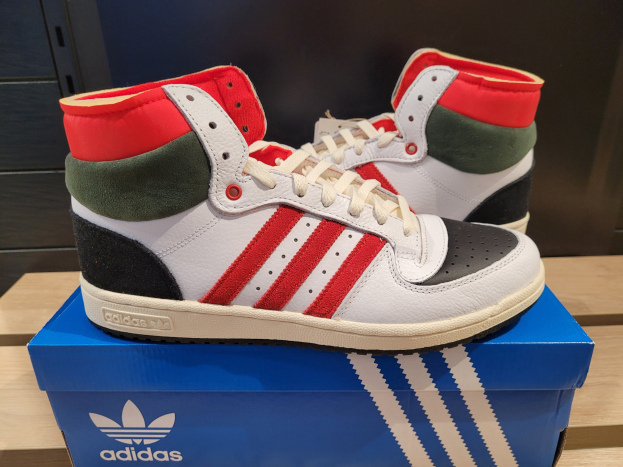 Weiße, rote, grüne und schwarze Adidas Originals Top Ten Hi Schuhe auf einer blauen und braunen Kartonschachtel, die auf einem cremefarbenen Tisch mit einer schwarzen Wand im Hintergrund steht.