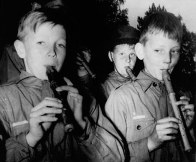 Eine Gruppe junger Jungs, die Flöten im Freien spielen, mit Bäumen im Hintergrund, in Schwarz-Weiß.