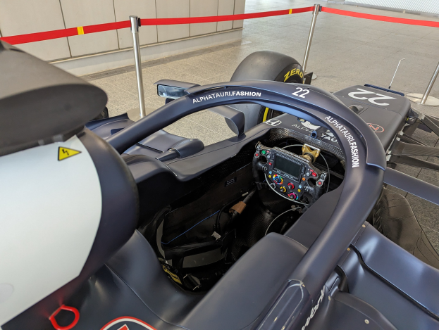 Cockpit eines Formel-E-Autos in einer Ausstellungshalle mit einem Lenkrad im Vordergrund, einer Wand im Hintergrund und dekorativen Bändern an Pfosten um das Fahrzeug.