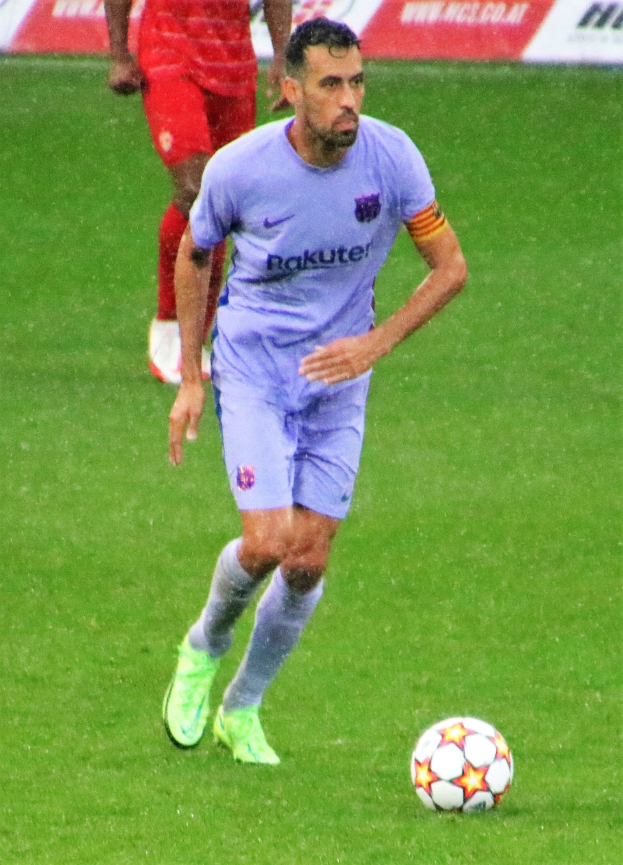 Fußballspieler in einem Sportoutfit und -schuhen auf einem Feld mit einer Texttafel im Hintergrund.