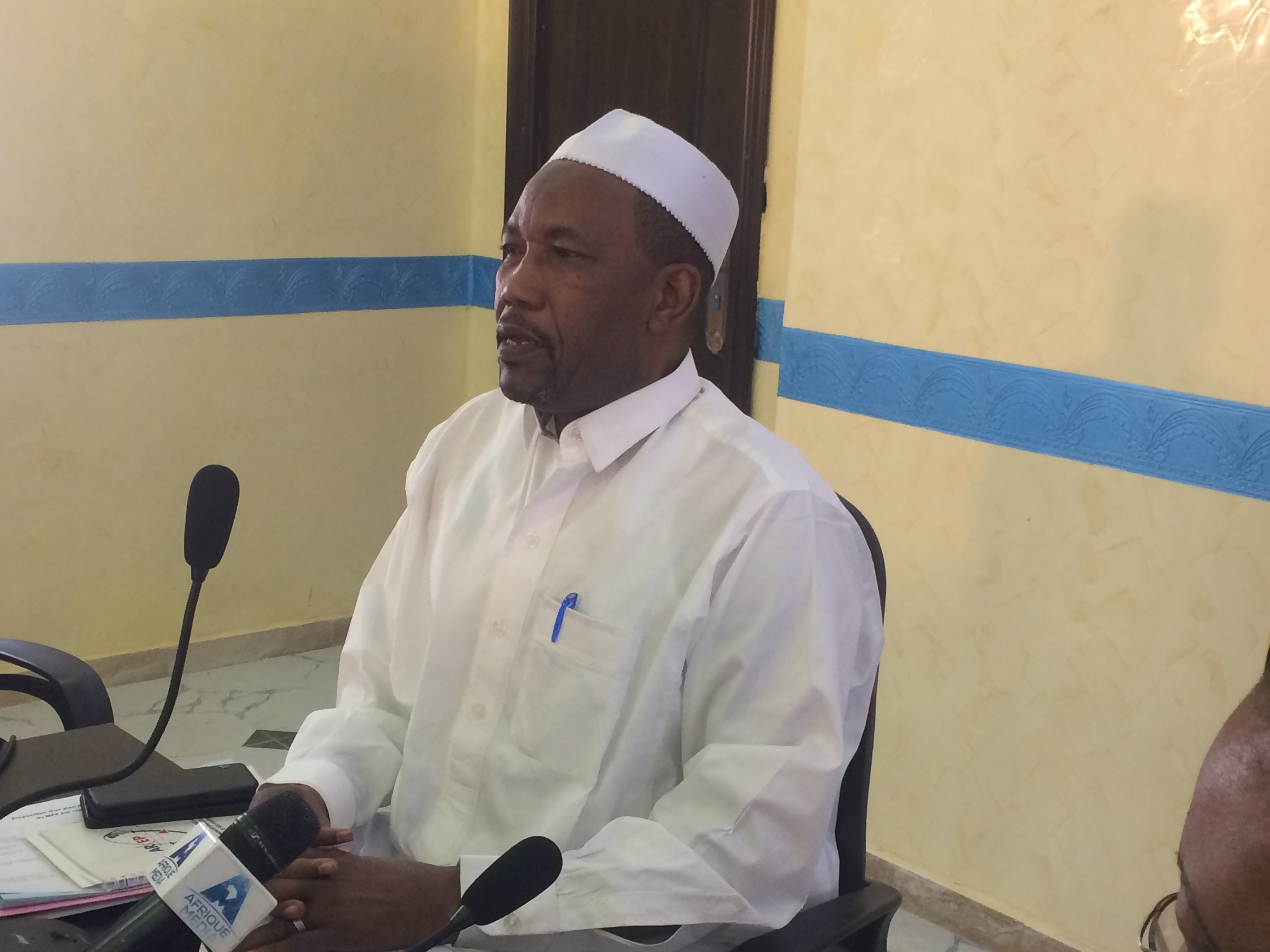 Ein Mann in einem weißen Gewand und einer Mütze sitzt an einem Tisch mit Mikrofonen und spricht, während Papiere und Gegenstände auf dem Tisch zu sehen sind, mit einem anderen Menschen mit Brille rechts von ihm, in einem Raum mit einer Wand und einer Tür im Hintergrund.