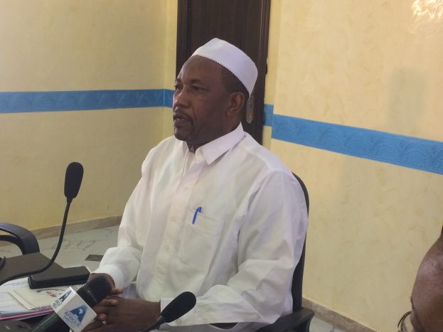 Ein Mann in einem weißen Gewand und einer Mütze sitzt an einem Tisch mit Mikrofonen und spricht, während Papiere und Gegenstände auf dem Tisch zu sehen sind, mit einem anderen Menschen mit Brille rechts von ihm, in einem Raum mit einer Wand und einer Tür im Hintergrund.