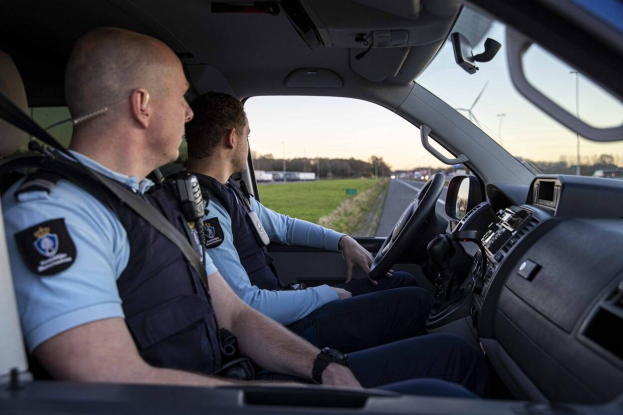 Zwei Polizeibeamte sitzen in einem Auto auf einer Straße mit Gras, Bäumen und Pfählen unter einem klaren blauen Himmel.