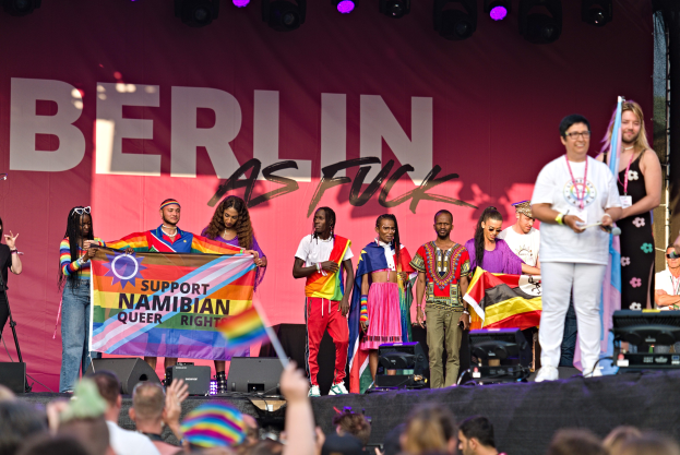 Gruppe von Menschen auf einer Bühne mit einem Banner, auf dem "Berlin as Fuck" steht, mit Rednern, Mikrofonen und einer feiernden Menge unten, die Hände hebt.