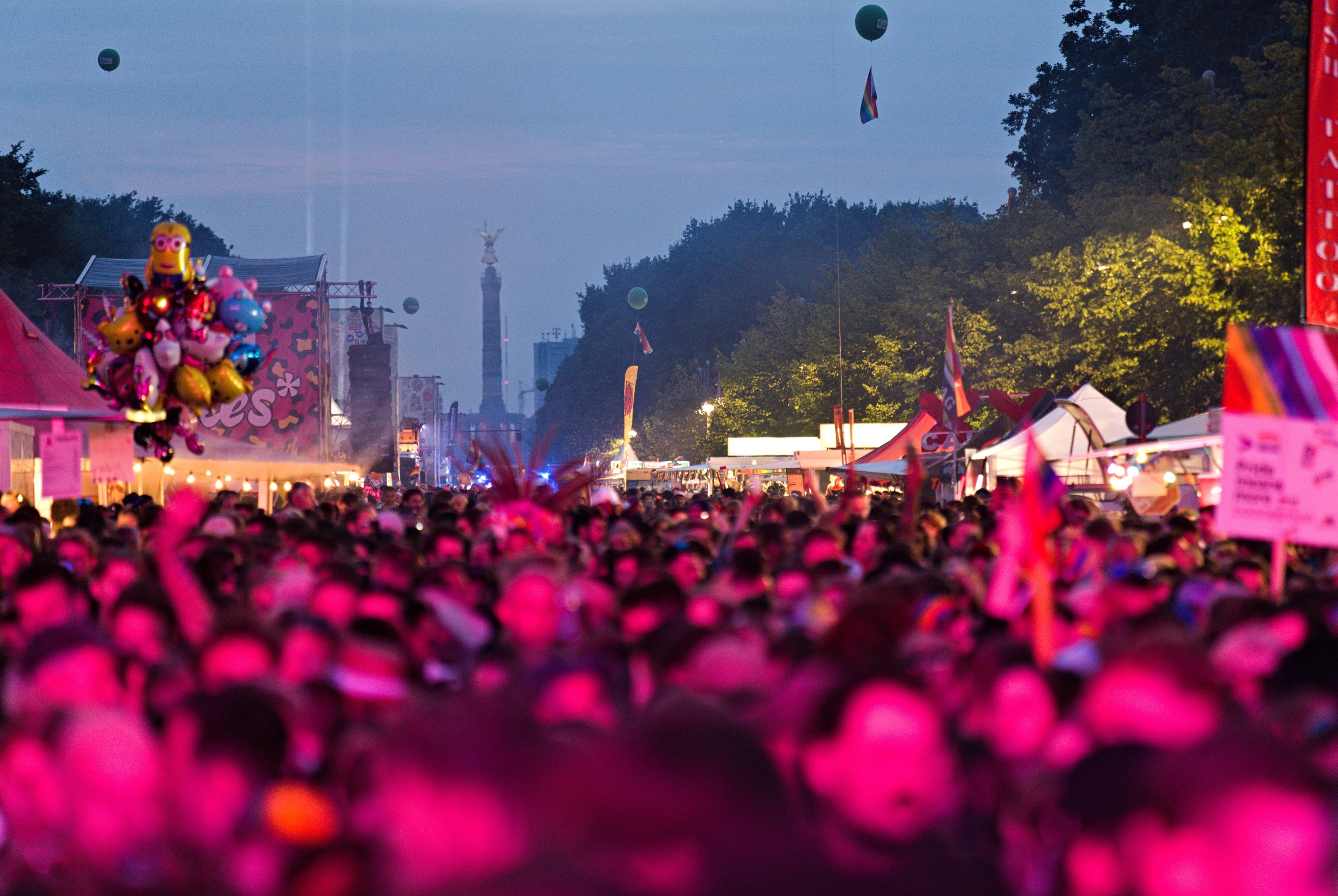 Eine große Menschenmenge auf einem nächtlichen Musikfestival mit bunten Lichtern, Ballons, Zelten, Bannern, Fahnen, Bäumen, einem Turm und einem sternenklaren Himmel im Hintergrund.
