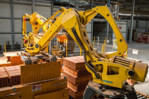 Ein großes Lagerhaus voller numerous boxes, Kräne und verschiedene industrielle Ausrüstung, mit einem Zaun und Pfählen im Vordergrund und einer Wand im Hintergrund.