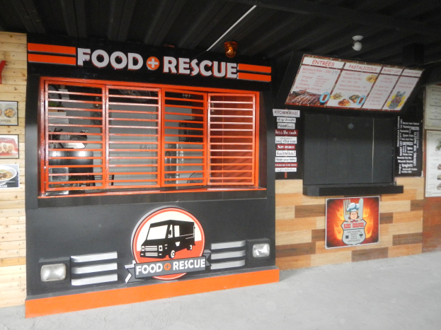 Ein Essenswagen steht vor einem Geb├Ąude, das mit "Food Rescue" beschriftet ist, mit Werbetafeln an der Wand und Deckenleuchten.