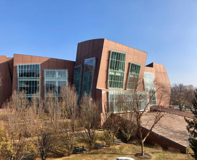 Großes Gebäude mit vielen Fenstern, umgeben von Bäumen, Bänken, Gras und einem Laternenmast, vor einem klaren blauen Himmel mit anderen Gebäuden im Hintergrund, als der neue Campus der University of Illinois at Chicago identifiziert.