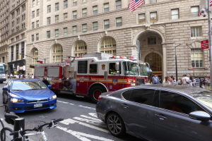 Ein roter Feuerwehrauto auf einer New Yorker Stra├če geparkt mit Fahrzeugen, Fü├čg├Ąngern, Fahrr├Ądern, Geb├Ąuden, Laternen und Flaggenmasten im Hintergrund.