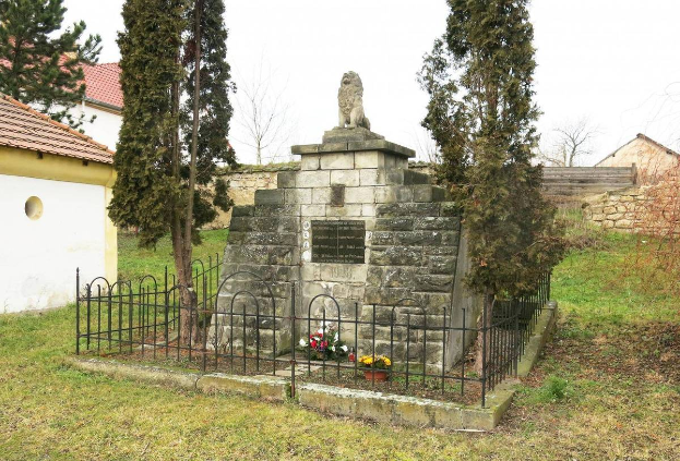 Ein steinernes Denkmal auf einer Wiese, umgeben von einem Metallzaun und geschmückt mit Blumensträußen, mit Bäumen, Häusern und einer Wand im Hintergrund unter einem klaren blauen Himmel dient als Holocaust-Gedenkstätte in Sibiu, Rumänien.